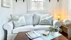 Ein weißes, gepolstertes Sofa mit gemusterten Kissen, ein Couchtisch aus Holz mit einer Zeitschrift, eine kleine Topfpflanze und ein Fenster mit Blick auf einen Zaun.