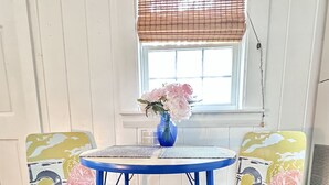 Ein kleiner, runder Tisch mit blauer Basis und weißer Tischplatte, begleitet von zwei Stühlen mit farbenfroher Blütenmuster-Polsterung. Auf dem Tisch steht eine Vase mit rosa Blumen.