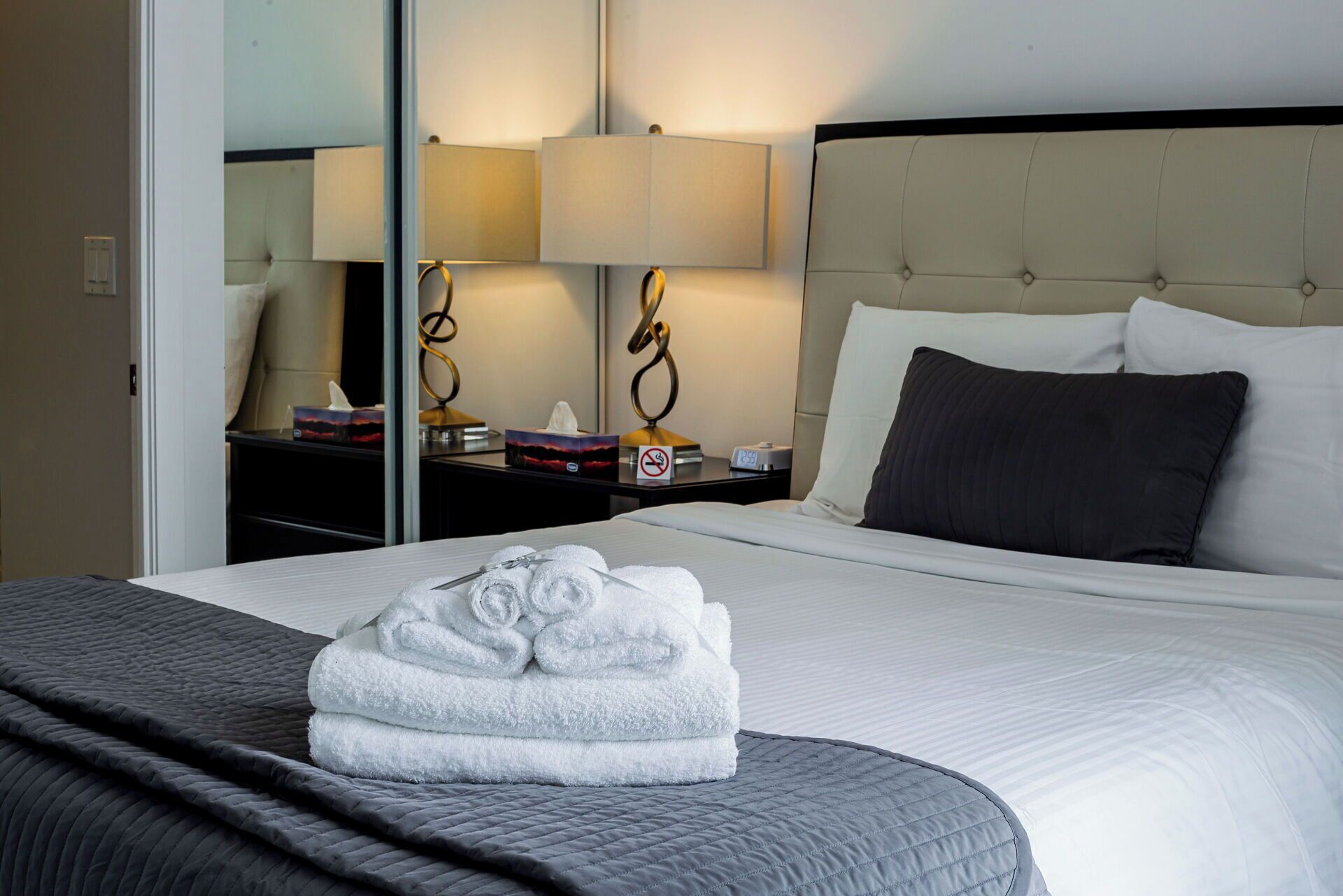 A neatly made bed with a grey quilt, a stack of white towels, a bedside lamp, and a nightstand with a clock and a no-smoking sign.