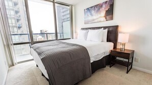 Un dormitorio moderno con una cama grande, una mesita de noche, una lámpara y vistas a la ciudad a través de ventanales del suelo al techo.
