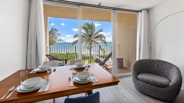 A dining area with a table set for four, a grey armchair, and a balcony with a view of the beach and palm trees.