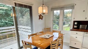 A dining area with a wooden table and chairs, a pendant light, and a view of the outdoors through large windows. - Jonathan Livingston Seagull:  Dog friendly single story home. (Ocracoke)