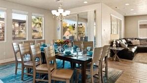 A dining area with a large table set for six, surrounded by chairs. There is a chandelier above the table, and the room has large windows with blinds. - PVZ 33|Steps to Pools *Misters* Pool/Mtn PatioView (Santa Clara)