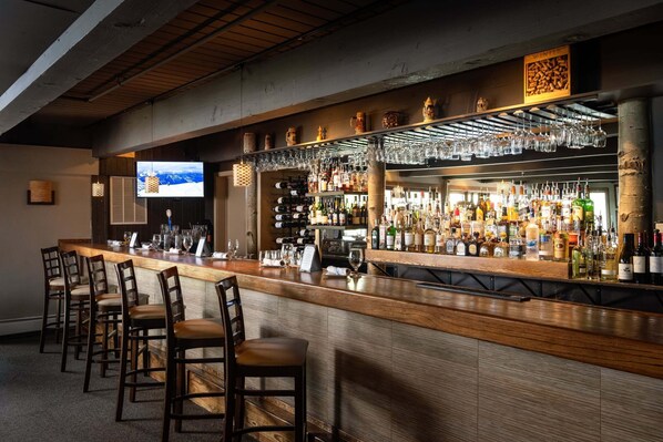 A bar area with a long counter, multiple bar stools, a television screen, and a variety of bottles and glasses.