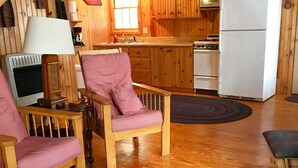 A cozy cabin interior with wooden walls, a kitchen area, a dining table with chairs, and a large woven rug. - Family Friendly Updated Waterfront Cottage (Leeds)