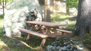 A picnic table in a forested area with a rustic wooden structure in the background. - Family Friendly Updated Waterfront Cottage (Leeds)