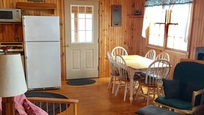 A wooden cabin interior with a dining table, chairs, a refrigerator, and a window with curtains. - Family Friendly Updated Waterfront Cottage (Leeds)