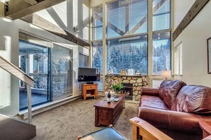 A living room with a stone fireplace, large windows, a leather sofa, and a television on a stand.