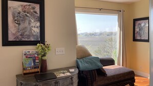 A room with a large window, a framed lion painting, a travel-themed bookcase, and a cushioned bench. - Folly Beach Riverfront Apt with dock, private boat ramp kayaks and bikes (Folly Beach)
