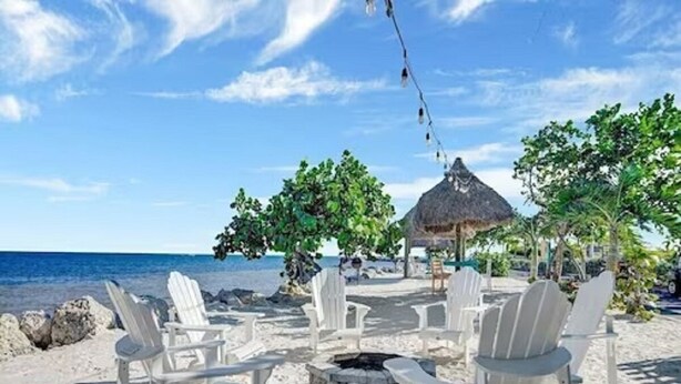 A beach setting with white lounge chairs arranged around a central fire pit, overlooking the ocean. - NEW! Casa Azul - NO HIDDEN FEES! Golf Cart, 2 Kings, Pool, Beach, Tennis, Kayaks (Key Largo)