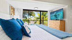 A bedroom with a large bed, a TV, and a balcony with a view of palm trees. - NEW! Casa Azul - NO HIDDEN FEES! Golf Cart, 2 Kings, Pool, Beach, Tennis, Kayaks (Key Largo)