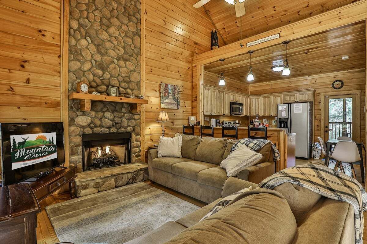 A cozy cabin interior with a stone fireplace, wooden walls, a living room with a sofa and a television, and a kitchen area.