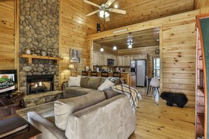 A cozy cabin interior with a stone fireplace, wooden walls, a large sofa, and a kitchen area.