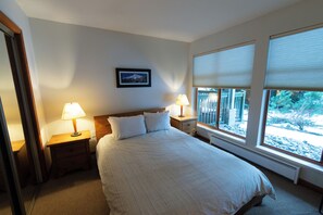 Un lit bien fait dans une chambre d’hôtel, avec deux tables de chevet, chacune équipée d’une lampe. Un tableau encadré orne le mur, et de grandes fenêtres sont dotées de stores.