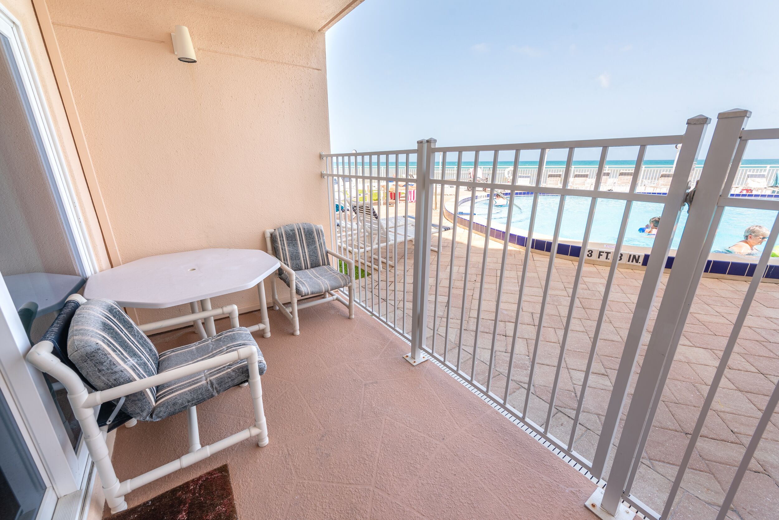 Un balcon avec une table blanche et deux chaises, une piscine dont la profondeur est indiquée à 3 pieds et 3 pouces, et une vue sur la plage.