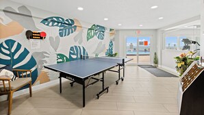 A ping pong table in a room with a leaf-patterned wall, a wooden chair, and a reception desk with a plant. - Steps from Pool & Beach, Oceanfront ground floor condo, Unit 106 (Satellite Beach)