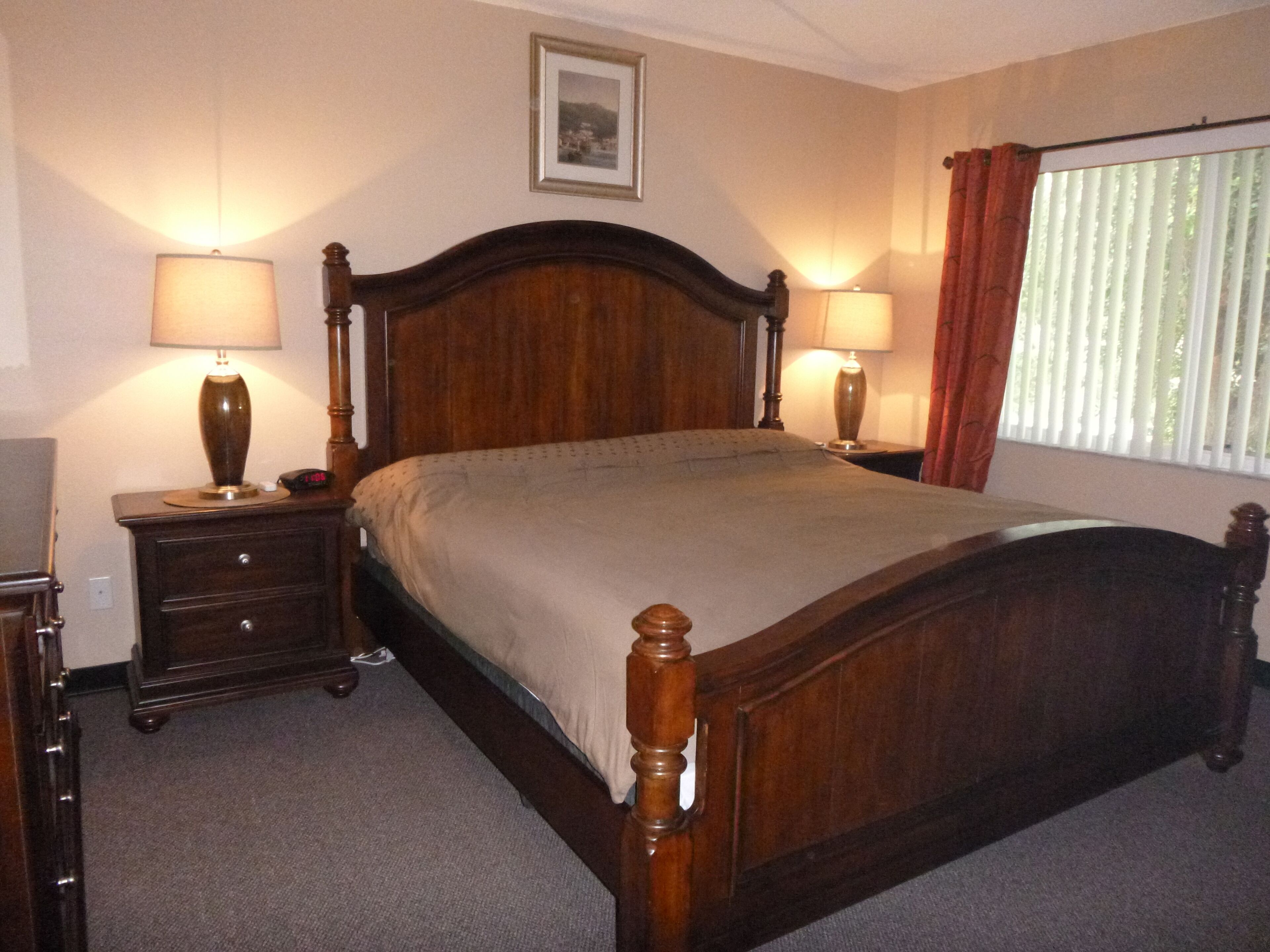 Un dormitorio ordenado con una cama de madera, dos mesitas de noche con lámparas, un cuadro en la pared y una ventana con cortinas.