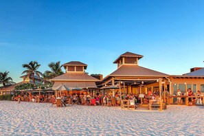 A beachfront resort with multiple dining areas and outdoor seating, surrounded by palm trees and a sandy beach. - Shorewalk rental By Owner/Condo 2bed/2bath/IMG Academy /Anna Maria Island (Bradenton)