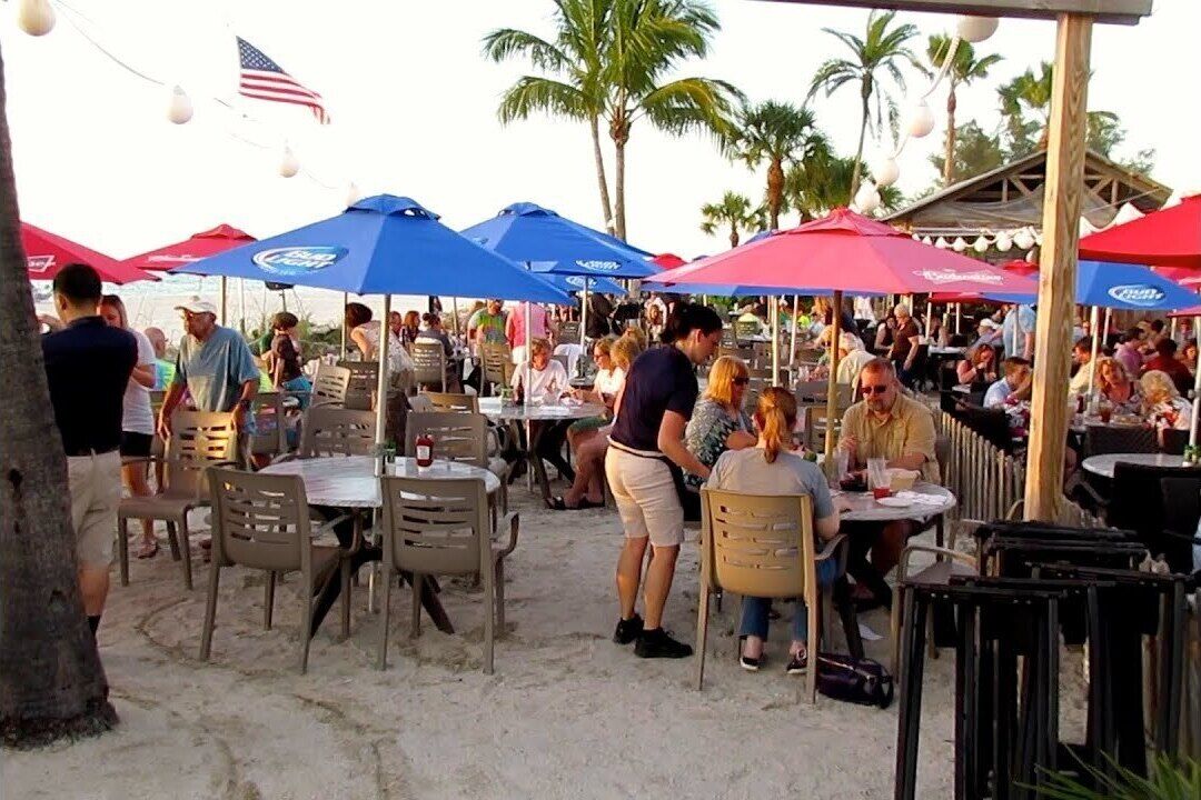 An outdoor dining area with tables and chairs, covered by umbrellas, and palm trees in the background.
