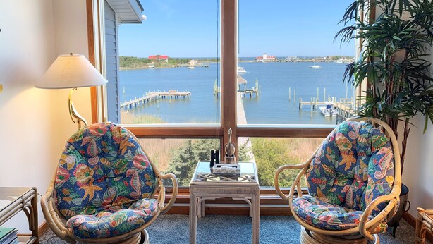 A room with two wicker chairs facing a large window overlooking a marina with boats and a lighthouse. - Pirate's Quay 3 North: Incredible harbor views and sunsets. Docking. (Ocracoke)