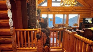 Ein Holzblockhaus-Innenraum mit einer Holztreppe, einer Kristallleuchter, einem Kamin aus Stein und einem Fernseher im Wohnbereich.