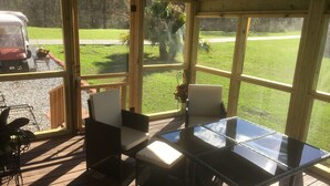 Un patio coperto con un tavolo e sedie in vetro, un’area esterna in legno e vista su una zona verde con un veicolo sullo sfondo.