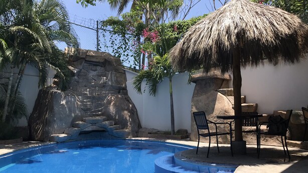 Una piscina con una cascada de piedra, rodeada por un muro blanco y palmeras.