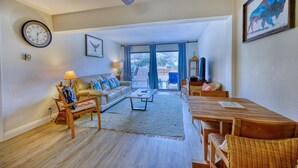 Ein geräumiges Wohnzimmer mit Sofa, Couchtisch und Esstisch. An der Wand hängt eine Uhr, und es gibt ein Fenster mit Vorhängen.