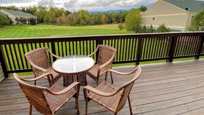 Una terraza de madera con sillas de mimbre y una mesa de vidrio, con vista a un césped cuidado y un edificio al fondo.