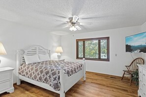 A bedroom with a bed, two bedside tables, a ceiling fan, a window with blinds, and a wall-mounted TV.