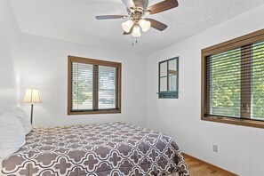 A bedroom with a bed, a ceiling fan, two windows with blinds, and a bedside lamp.