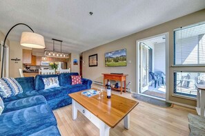 A living room with a blue sofa, a wooden coffee table, and a kitchen in the background. - Greystone Lodge 202 Ski-in/Out by Acer Vacations (Whistler)