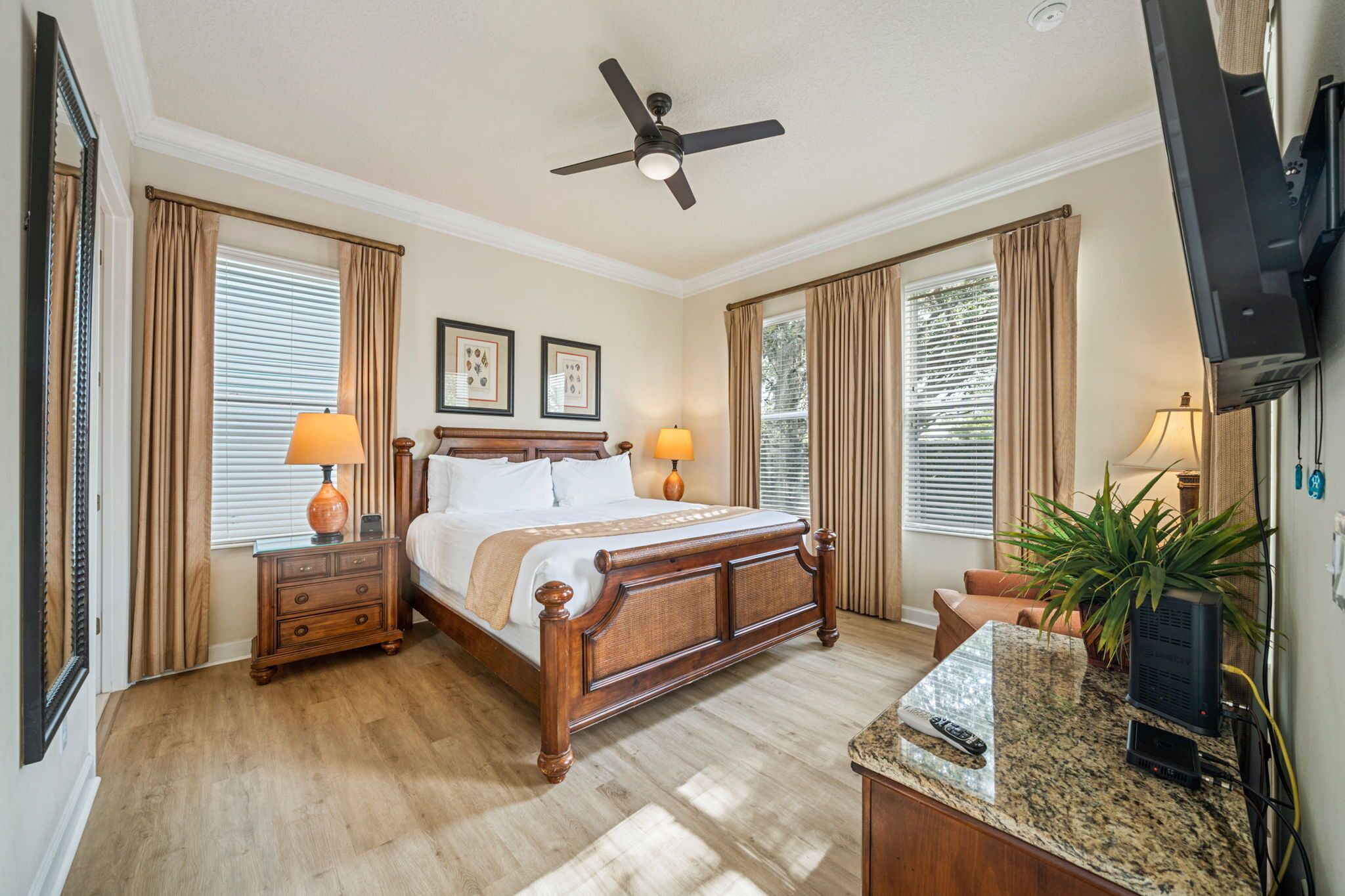 A hotel room with a wooden bed, two bedside tables with lamps, a ceiling fan, a television, and a large window with curtains.