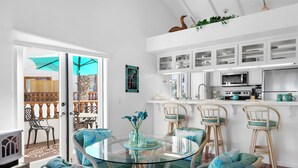 A dining area with a glass table surrounded by wicker chairs, a kitchen with white cabinets, and a view of a balcony with wooden furniture. - H962: Great Ocean Views, Golf Cart + Only 10 Steps + King Bed + Fireplace (Avalon)