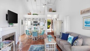 A spacious living area with a fireplace, a sofa, and a television. There is a dining area with chairs and a table. The kitchen is visible in the background. - H962: Great Ocean Views, Golf Cart + Only 10 Steps + King Bed + Fireplace (Avalon)