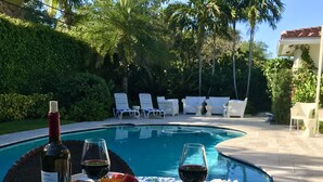 A poolside dining setup with a table, plates of food, and wine glasses. - Luxury East Boca Home • Beautiful Tropical Pool / Patio Oasis • Perfect Location (Boca Raton)