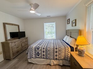 Une chambre à coucher comprenant un lit, une commode, un téléviseur, un ventilateur de plafond, une fenêtre avec des stores et une lampe de chevet.
