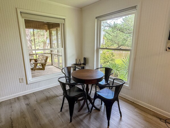 A room with a round wooden table and four chairs, a window with a view of greenery, and a porch with a swing.