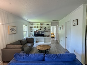 A living room with a sofa, a coffee table, and a TV. There is a bookshelf with various items and a chair in the background.