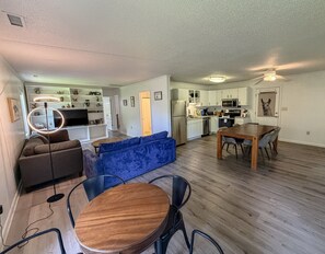 A spacious living area with a blue sofa, a wooden dining table, and a kitchen in the background.