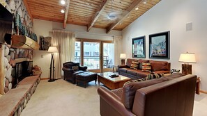 A cozy living room with a stone fireplace, leather sofas, and a large window with curtains. - 🌲Timberline B3H Deluxe 2-Bedroom + Loft ~ 3/bath ~Hot Tub~Pool~WiFi~Ski-in/out~Local Shuttle🌲 (Snowmass Village)