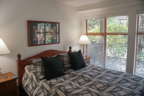 A bedroom with a bed, two bedside tables, a nightstand, a window with blinds, and a framed artwork on the wall.
