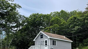 A single-story house with a front porch and a small deck, surrounded by a grassy yard and dense trees. - Camp Forever cottage (Harpswell)
