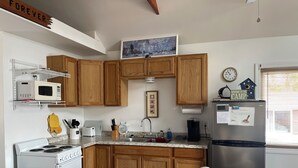 A kitchen with wooden cabinets, a stainless steel refrigerator, a microwave, a stove, and a sink. There is a table with magazines and a pair of scissors. A clock and a welcome sign are on the wall. - Camp Forever cottage (Harpswell)