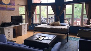 Un dormitorio con cama, un escritorio con televisor, un sofá y ventanas amplias con cortinas.