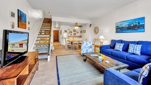 Ein geräumiges Wohnzimmer mit einem blauen Sofa, einer Holz-Unterhaltungseinheit und einem Fernseher. Eine Treppe führt zu einem Essbereich mit Tisch und Stühlen.