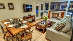 A dining area with a wooden table and chairs, a sofa, a fireplace, and a television mounted on the wall. - Greystone Lodge 109 Ski-in/Out by Acer Vacations (Whistler)