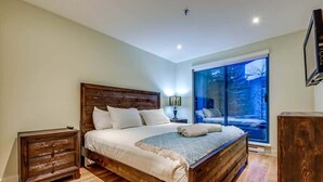 A bedroom with a wooden bed, a nightstand, a lamp, and a large window with a view. - Greystone Lodge 109 Ski-in/Out by Acer Vacations (Whistler)