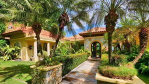 A courtyard with a stone pathway leading to a building with arched doorways, surrounded by lush greenery and palm trees. - Spectacular Hilltop Rental for 2 w/ Private Beach, Mountain & Sea Views. (Roatan)