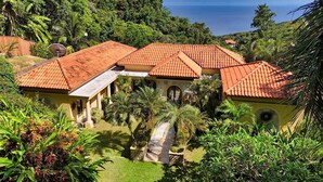 A villa with a red-tiled roof, surrounded by lush greenery and a view of the ocean. - Spectacular Hilltop Rental for 2 w/ Private Beach, Mountain & Sea Views. (Roatan)